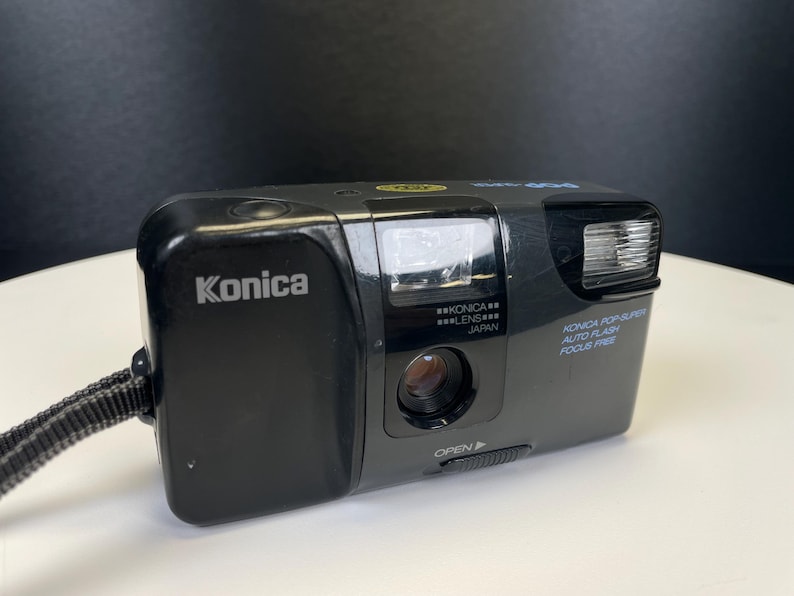May include: Black Konica Pop Super 35mm film camera with a built-in flash. The camera has the brand name "Konica" printed on the front. The camera has a lens and the words "AUTO FLASH FOCUS FREE" are printed on the side.