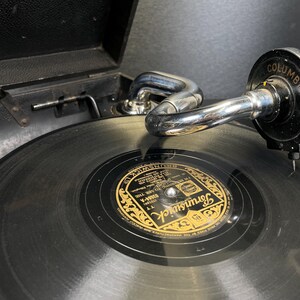Puede incluir: Primer plano de un tocadiscos vintage con un disco de vinilo negro. La etiqueta del disco es dorada y negra, con la palabra "Brunswick" visible. El brazo fonocaptor es cromado y el cabezal es negro con la palabra "Columbia".