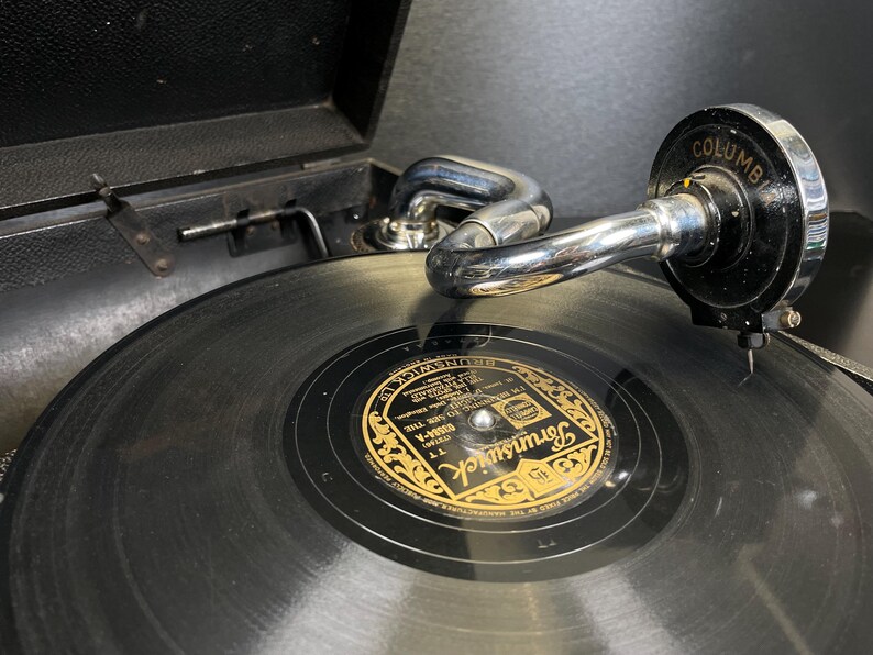 Puede incluir: Primer plano de un tocadiscos vintage con un disco de vinilo negro. La etiqueta del disco es dorada y negra, con la palabra "Brunswick" visible. El brazo fonocaptor es cromado y el cabezal es negro con la palabra "Columbia".