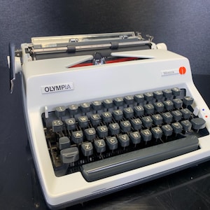 Può includere: Una macchina da scrivere Olympia Monica vintage bianca con tasti neri. La macchina da scrivere ha il marchio "OLYMPIA" sul davanti e il nome del modello "Monica" in alto a destra. I tasti sono disposti in un layout QWERTY.