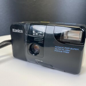 May include: Black Konica Pop-Super camera with auto flash and focus-free features. The camera has a lens and flash on the front, with the Konica brand name on the top left. The camera has a wrist strap.