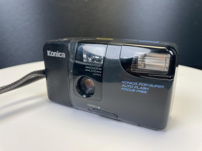 May include: Black Konica Pop-Super camera with auto flash and focus-free features. The camera has a lens and flash on the front, with the Konica brand name on the top left. The camera has a wrist strap.