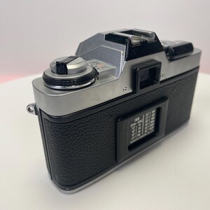 May include: A silver and black 35mm film camera with a black leatherette covering. The camera has a silver lens and a black viewfinder. The camera has a silver dial on the top and a black dial on the side.