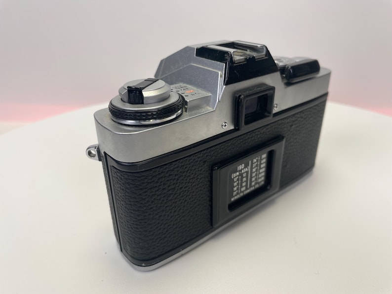 May include: A silver and black 35mm film camera with a black leatherette covering. The camera has a silver lens and a black viewfinder. The camera has a silver dial on the top and a black dial on the side.