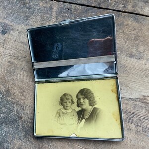 Könnte beinhalten: Ein silbernes Zigarettenetui aus Metall mit einem Foto von zwei Personen im Inneren. Das Foto zeigt eine Frau mit dunklen Haaren und ein Mädchen mit hellen Haaren. Die Frau trägt ein weißes Kleid und das Mädchen trägt ein weißes Kleid mit einer roten Schleife. Der Text "Ora et Labora" ist auf dem Etui aufgedruckt.