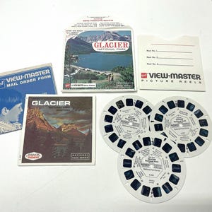 Op de afbeelding: Drie vintage View-Master-rollen met afbeeldingen van Glacier National Park in Montana. De rollen zijn wit met blauwe en zwarte afbeeldingen. De rollen zijn gelabeld met "See-Thru Glacier National Park".