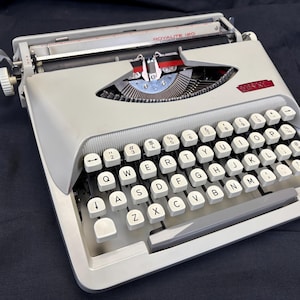 Puede incluir: Una máquina de escribir Royalite 120 vintage en gris claro. La máquina de escribir tiene un teclado QWERTY completo con teclas blancas y letras negras. La palanca de retorno del carro y otros detalles son cromados.