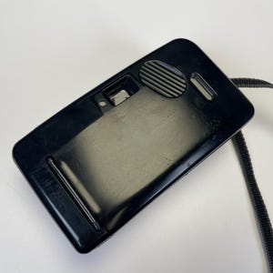 May include: A black, rectangular camera with a strap. The camera has a glossy finish and a few small details, including a circular dial and a small lens. The strap is black and made of a woven material.