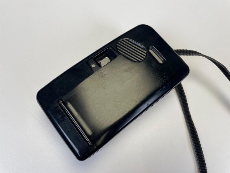 May include: A black, rectangular camera with a strap. The camera has a glossy finish and a few small details, including a circular dial and a small lens. The strap is black and made of a woven material.