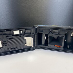 May include: Black film camera with the back open, revealing the interior components. The camera has a strap and is on a white surface. The camera is labeled "POP SUPER".