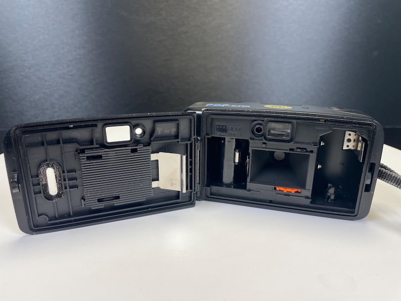 May include: Black film camera with the back open, revealing the interior components. The camera has a strap and is on a white surface. The camera is labeled "POP SUPER".