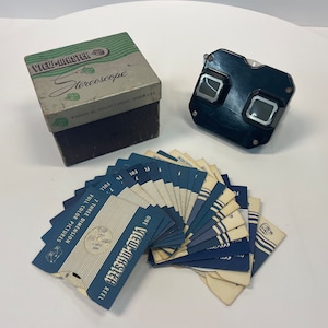Op de afbeelding: Vintage View-Master Stereoscoop set met een zwarte viewer, een doos en verschillende blauw-witte reels. De doos is grijs met groene accenten en de woorden "View-Master Stereoscope" staan erop. De reels zijn gelabeld met "Full Color Pictures".