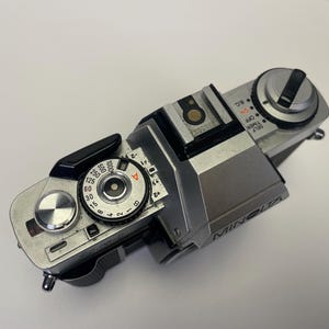 May include: A silver Minolta film camera with a black leather grip. The camera has a dial for setting the shutter speed and a dial for setting the aperture. The camera also has a self-timer and a timer.