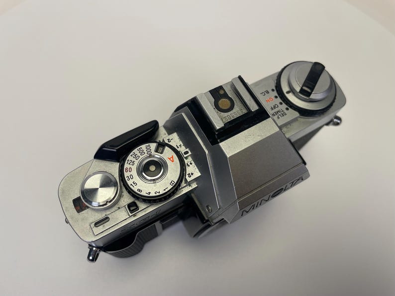 May include: A silver Minolta film camera with a black leather grip. The camera has a dial for setting the shutter speed and a dial for setting the aperture. The camera also has a self-timer and a timer.
