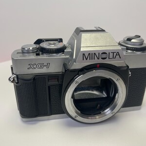 May include: A silver Minolta XG-1 film camera with a black leather grip and a silver lens. The camera has a black and silver body with a silver lens.