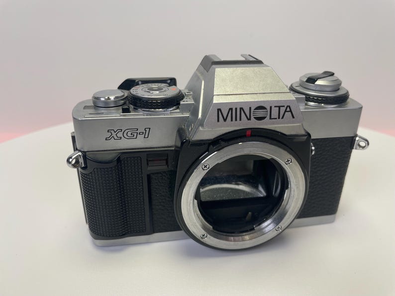 May include: A silver Minolta XG-1 film camera with a black leather grip and a silver lens. The camera has a black and silver body with a silver lens.