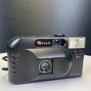 Könnte beinhalten: Schwarze Fuji-Filmkamera mit Fujinon-Objektiv. Die Kamera verfügt über einen eingebauten Blitz und eine Drop-in-Ladefunktion. Sie hat einen schwarzen Riemen und goldfarbene Schrift.