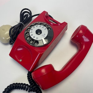 Op de afbeelding: Een rode draaischijftelefoon met een zwart snoer en een witte draaischijf. De telefoon heeft een wit label met de tekst "SATURN" en "MADE IN ITALY".