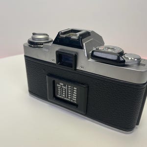 May include: A silver and black 35mm film camera with a black leatherette covering. The camera has a silver lens and a black film advance lever. The camera has a black film speed dial with the text "ISO (DIN - ASA)" and a range of numbers from 12 to 3200.