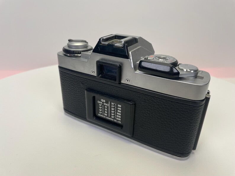 May include: A silver and black 35mm film camera with a black leatherette covering. The camera has a silver lens and a black film advance lever. The camera has a black film speed dial with the text "ISO (DIN - ASA)" and a range of numbers from 12 to 3200.