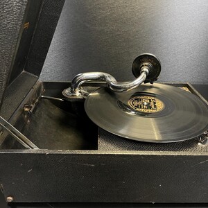 Puede incluir: Un tocadiscos Columbia antiguo en una caja negra con un brazo plateado y un disco negro en el tocadiscos. El tocadiscos est&aacute; abierto, revelando el interior. El disco tiene una etiqueta dorada y negra.