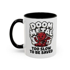 May include: A white ceramic mug with a black handle and interior rim. The front features a graphic of a red devil character with wings, holding an axe and making a hand gesture, surrounded by clouds. Text reads DOOM METAL and TOO SLOW TO BE SAVED.
