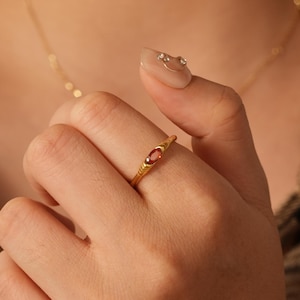May include: A gold ring featuring a small, oval red gemstone. The ring has a textured band and is worn on a finger. A gold necklace is also visible. The background is a plaid shirt.