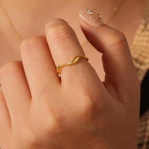 Könnte beinhalten: Ein goldfarbener Ring mit einem wellenförmigen Design. Der Ring wird an einem Finger getragen. Der Hintergrund umfasst eine goldene Halskette und ein kariertes Hemd.