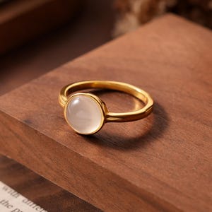 May include: A gold-colored ring with a round, milky-white stone set in the center. The ring is displayed on a dark wooden surface, showcasing its polished finish and elegant design. The ring's band is thin and delicate.