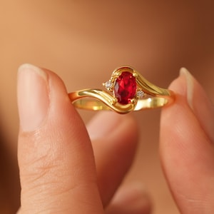 Può includere: Un anello d'oro con una vibrante gemma rossa ovale come elemento centrale, affiancata da due gemme bianche più piccole. La fascia dell'anello ha un design fluido ed elegante. L'anello è tenuto dalle dita di una persona.