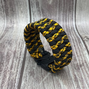 May include: A black and yellow paracord bracelet with a black plastic clasp. The bracelet is made of tightly woven cord in a repeating pattern of black and yellow. The bracelet is shown on a wooden surface.