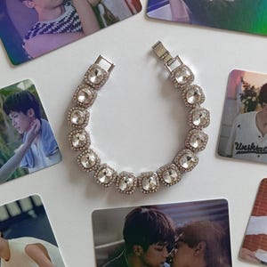 Può includere: Un braccialetto d'argento con pietre preziose quadrate e trasparenti, ciascuna circondata da pietre più piccole e trasparenti. L'immagine include anche diverse piccole carte con immagini di persone.