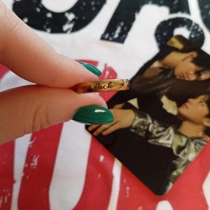 Puede incluir: Un anillo dorado con el nombre "Jack" grabado. El anillo está sostenido por una mano con esmalte de uñas verde. En el fondo, una fotografía de dos personas.