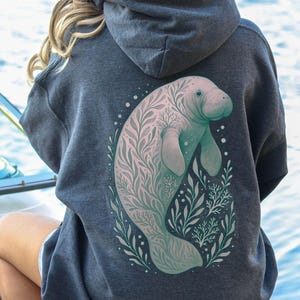 May include: A dark gray hoodie with a large illustration of a manatee on the back. The manatee is light blue and pink, surrounded by green and white foliage. The hoodie has a hood and is made of a soft material.