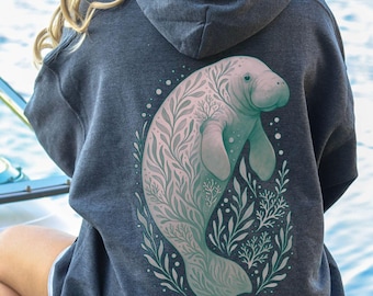 May include: A dark gray hoodie with a large illustration of a manatee on the back. The manatee is light blue and pink, surrounded by green and white foliage. The hoodie has a hood and is made of a soft material.