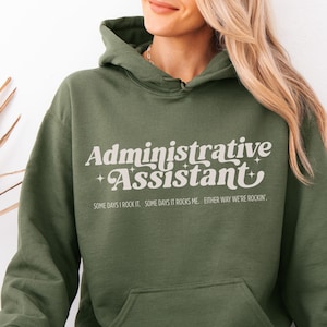 May include: An olive green hoodie featuring the text "Administrative Assistant" in a retro style. The text "SOME DAYS I ROCK IT. SOME DAYS IT ROCKS ME. EITHER WAY WE'RE ROCKIN'" is below. The hoodie has a front pocket and a hood.