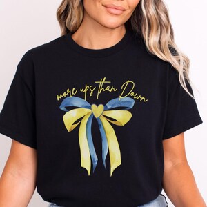 May include: Black t-shirt featuring a yellow and blue ribbon bow with the text "more ups than down" in gold lettering.