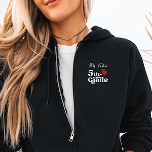 Può includere: Felpa nera con zip con la scritta "Ms. Katie 5th Grade" in corsivo bianco e una grafica di mela rossa. La felpa ha una cerniera completa e una maglietta bianca sotto.