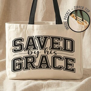 May include: Cream-colored tote bag with black handles and the text "SAVED by his GRACE" in a bold, collegiate-style font. Features a zipper top and an inside pocket. The bag is made of a sturdy canvas material.