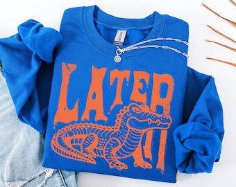 May include: Royal blue long-sleeve shirt with the word "LATER" in orange block letters and an orange alligator graphic. A silver necklace with a circular pendant rests on the shirt. The shirt is folded on a white surface.