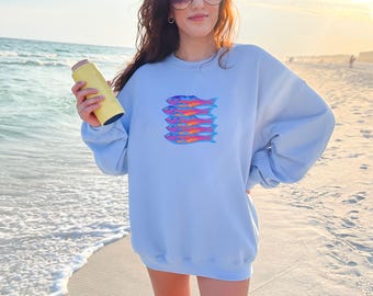 May include: Light blue sweatshirt featuring a stack of colorful fish. The fish are in shades of pink, orange, and blue. The sweatshirt is a relaxed fit and is being held by a person on a beach.