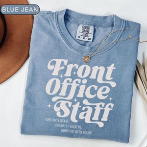 May include: A blue jean Comfort Colors t-shirt with the text "Front Office Staff" in a retro font. The shirt also has the phrases "Some days I rock it. Some days it rocks me. Either way we're rockin'." A gold necklace is draped across the shirt.