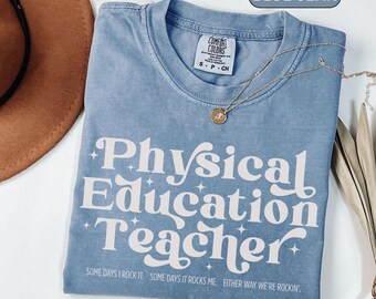 May include: A blue jean colored t-shirt with the words "Physical Education Teacher" in white, with star accents. The shirt also has the phrases "Some days I rock it. Some days it rocks me. Either way we're rockin'" printed on it. A gold necklace is draped across the shirt.