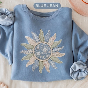 May include: A light blue crewneck sweatshirt featuring a large, central sun-shaped applique made from various fabric patches in shades of blue, green, and cream with floral and gingham patterns. The text BLUE JEAN is printed at the top.