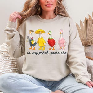 May include: Beige sweatshirt featuring a whimsical illustration of four geese dressed in various outfits. One goose holds a trowel, another wears a yellow raincoat, a third is a strawberry, and the last wears a pink robe. The text "in my porch goose era" is below the geese.