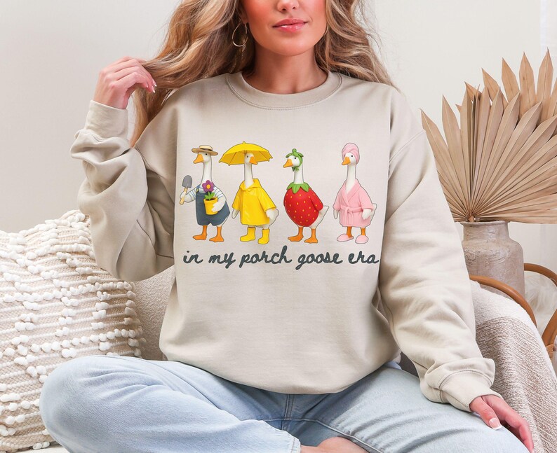 May include: Beige sweatshirt featuring a whimsical illustration of four geese dressed in various outfits. One goose holds a trowel, another wears a yellow raincoat, a third is a strawberry, and the last wears a pink robe. The text "in my porch goose era" is below the geese.