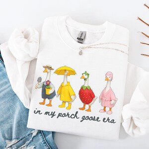May include: White sweatshirt featuring a whimsical illustration of four geese dressed in various outfits. One goose holds a trowel, another wears a yellow raincoat, a third is a strawberry, and the last wears a pink robe. The text "in my porch goose era" is below the geese.