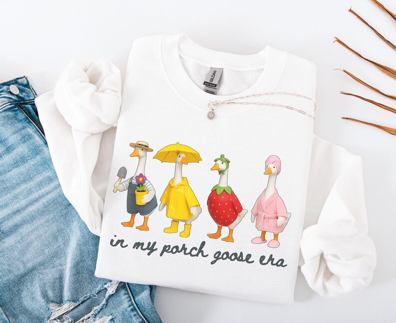 May include: White sweatshirt featuring a whimsical illustration of four geese dressed in various outfits. One goose holds a trowel, another wears a yellow raincoat, a third is a strawberry, and the last wears a pink robe. The text "in my porch goose era" is below the geese.