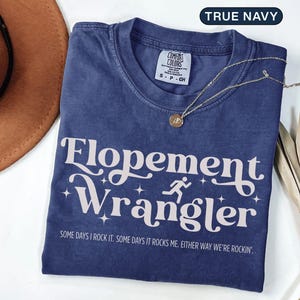 May include: A navy blue t-shirt with the text "Flopement Wrangler" in a white, stylized font. The shirt also features a running figure graphic and the phrase "SOME DAYS I ROCK IT. SOME DAYS IT ROCKS ME. EITHER WAY WE'RE ROCKIN'." A gold necklace rests on the shirt.