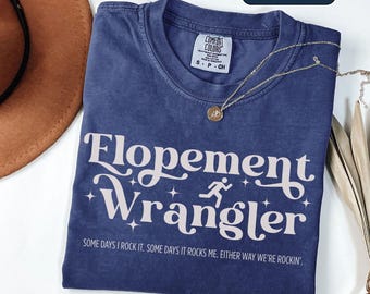 May include: A navy blue t-shirt with the text "Flopement Wrangler" in a white, stylized font. The shirt also features a running figure graphic and the phrase "SOME DAYS I ROCK IT. SOME DAYS IT ROCKS ME. EITHER WAY WE'RE ROCKIN'." A gold necklace rests on the shirt.